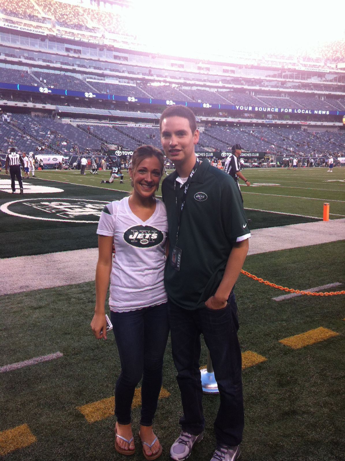 Justin Herrera in a Jets staff polo with a field credential, standing with Steph on the field at MetLife Stadium before kickoff.