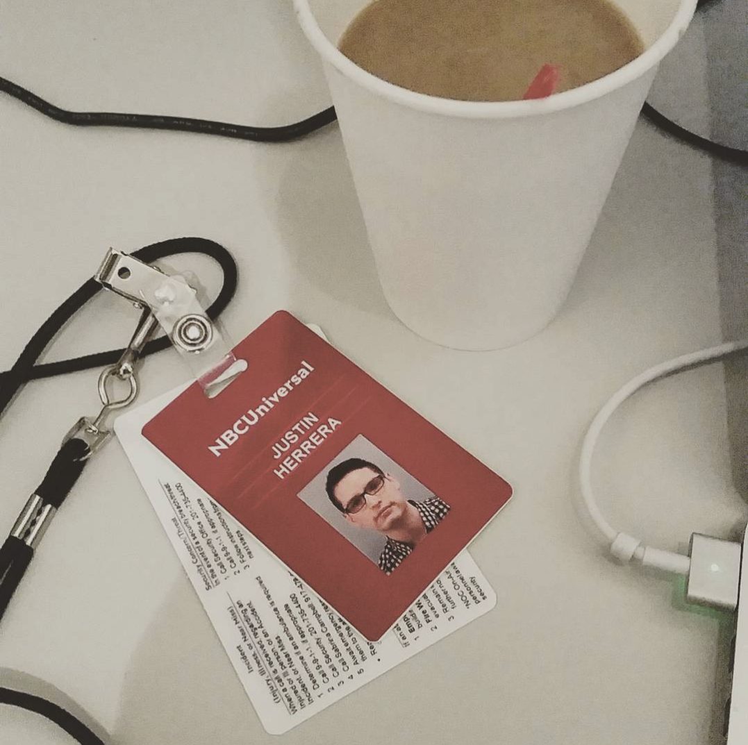 An NBCUniversal ID badge reading Justin Herrera, lying on a desk beside a coffee cup and a laptop charger.